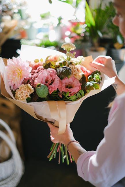 A person holding a beautifully arranged bouquet of fresh flowers, including large pink peonies, soft yellow and white carnations, and delicate purple and white blossoms, all nestled within lush green