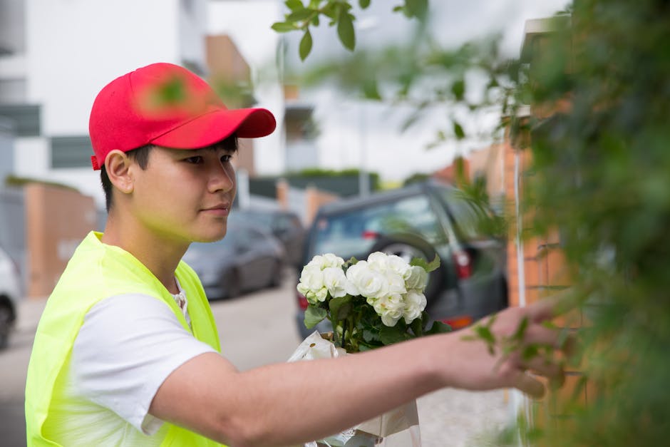 A young delivery person wearing a red cap and a neon yellow safety vest is standing outdoors on a residential street, holding a bouquet of fresh white roses wrapped in white paper with green foliage.
