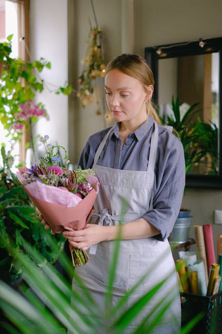 A young woman with light brown hair styled neatly, wearing a grey collared shirt and a light grey apron, is holding a bouquet of fresh flowers wrapped in pink paper. The bouquet features a mix of pink