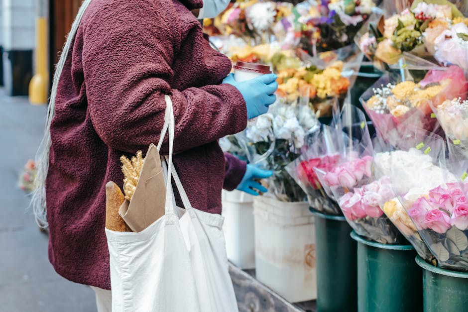 A person wearing a maroon fleece jacket and blue disposable gloves is shopping at an outdoor flower stall. They hold a white tote bag containing a paper bag and a small bundle of dried wheat stalks. T