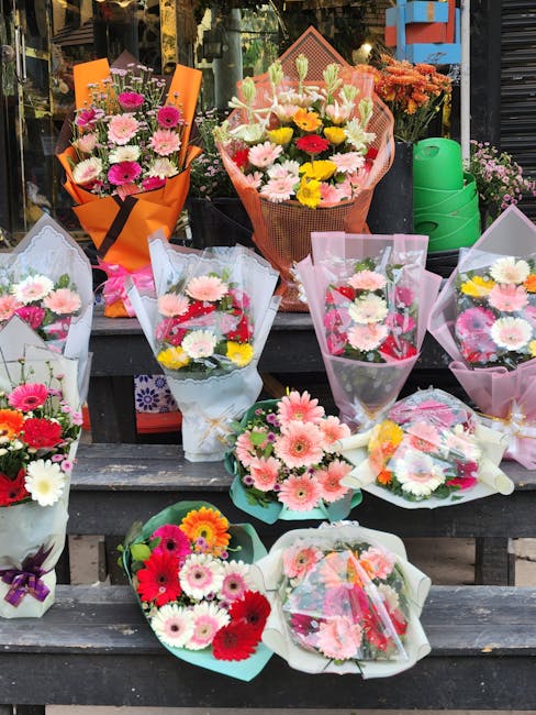 A display of various flower bouquets arranged on wooden benches outside a flower shop. The bouquets feature a mix of fresh, colorful flowers including pink, white, yellow, and red blooms such as gerbe