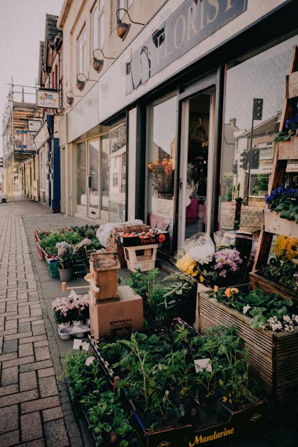 The image showcases the exterior of Hornchurch Flowers, a florist shop with large glass windows and a beige facade, displaying a vibrant array of fresh flowers and plants arranged on the sidewalk. In