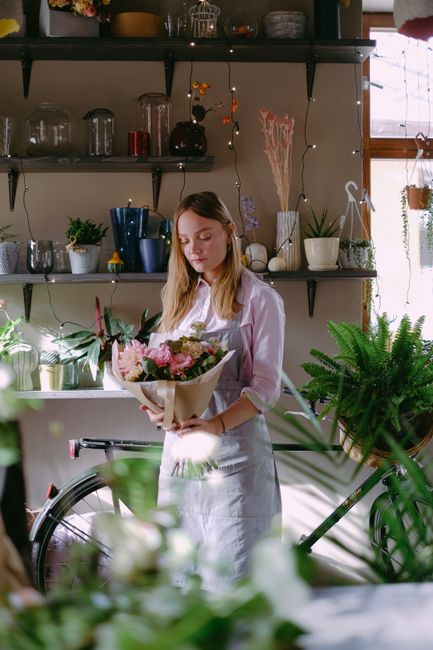 A young woman with light brown hair, dressed in a white apron and shirt, is holding a bouquet of fresh flowers wrapped in beige paper. The bouquet features pink roses, white lilies, and various green 
