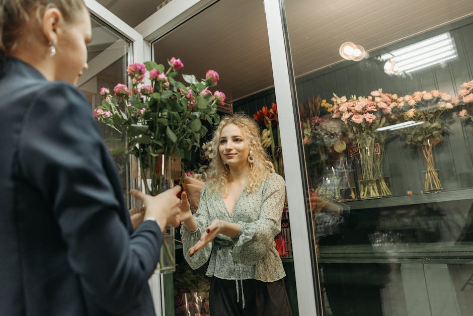A woman with curly blonde hair and light skin is inside a flower shop, receiving a bouquet of pink roses wrapped in brown paper from a florist. The woman is smiling gently and making a hand gesture, l